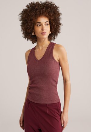 Femme aux cheveux bouclés portant des boucles d'oreilles anneaux en or, un haut en tricot sans manches de couleur bordeaux et un pantalon assorti, regardant sur le côté.