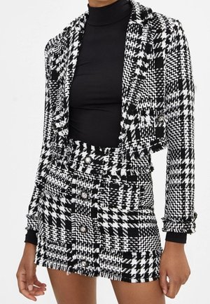 Zwart-witte blazer en rok met een pied-de-poule patroon. Gestructureerde stof met gerimpelde details en knoopsluitingen. Lange mouwen aan de blazer.
