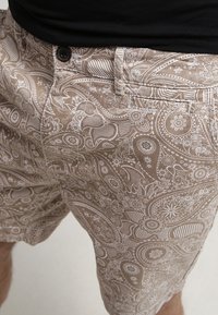 Shorts beiges avec des motifs paisley et floraux blancs complexes, dotés d'une fermeture à bouton et de poches latérales. Fabriqués en tissu léger.