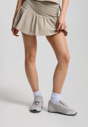Persona che indossa una gonna beige con volant, calzini bianchi e scarpe da ginnastica grigio chiaro, in piedi con le gambe leggermente divaricate su uno sfondo semplice.