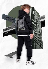 Melna kapuce ar grafisko dizainu, saskaņojamas melnas kravas bikses un baltas sporta apavu akcenti. Skaids atrodas blakus; pilsētas fons iezīmē arhitektūru.