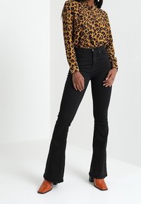 Camicia a stampa leopardata con bottoni, jeans a zampa neri e stivaletti arancioni con tacco. La camicia ha il colletto, i jeans sono a vita alta e presentano una texture liscia.