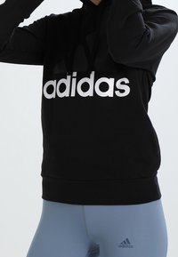 Person som bär en svart Adidas-hoodie med vit text och ljusblå leggings, stående mot en enfärgad bakgrund.