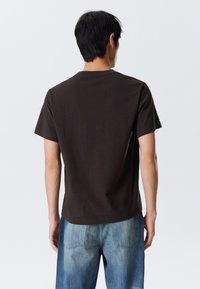 T-shirt à manches courtes marron foncé avec col rond, en matière de coton doux, présenté de dos avec un jean en denim bleu.