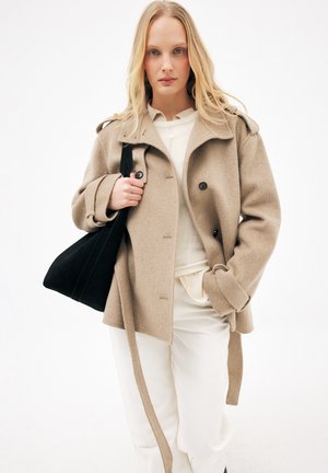 Blond naine kannab beežist villast mantlit valge riietuse peal, hoides musta õlakotti ühetaustalise valge tausta ees.