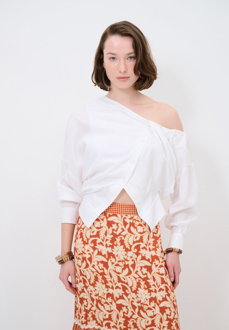 Blouse blanche dénudée des épaules avec des détails de boutons, associée à une jupe orange à motifs, ornée de motifs floraux et d'une ceinture à carreaux.