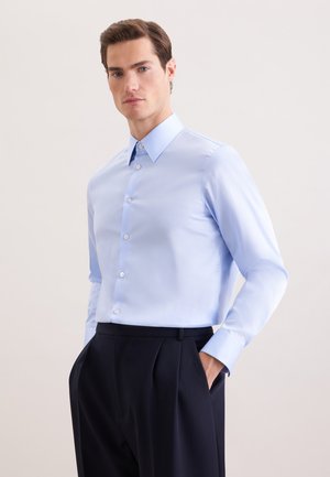 Homme portant une chemise bleu clair boutonnée, rentrée dans un pantalon noir plissé, avec une main dans la poche, sur un fond uni.