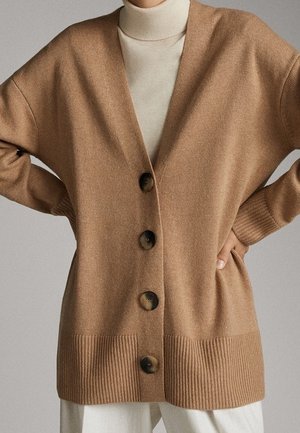 Person iført en kamel-farvet cardigan med knapper over en hvid højhalset trøje, med hænderne på hofterne og cremefarvede bukser.