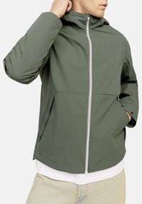 Giacca verde oliva con cappuccio, zip frontale e tasche laterali. Presenta un tessuto liscio e una vestibilità leggermente ampia, indossata sopra una maglietta bianca.