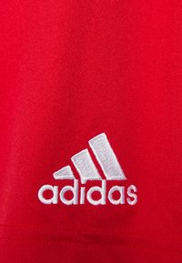 Rød sportstrøje lavet af struktureret stof, med et hvidt broderet Adidas-logo i nederste hjørne.