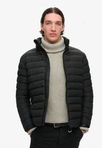 Sort pufferjakke med quiltet mønster, lynlåslukning og hætte. Båret over en lys grå ribbet turtleneck og sorte bukser.