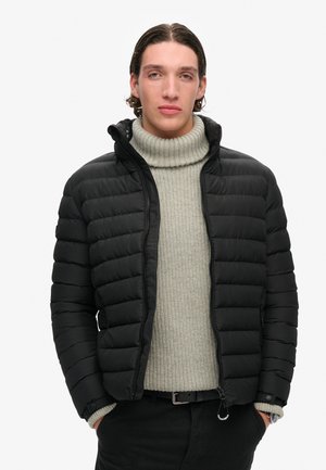 Sort pufferjakke med quiltet mønster, lynlåslukning og hætte. Båret over en lys grå ribbet turtleneck og sorte bukser.