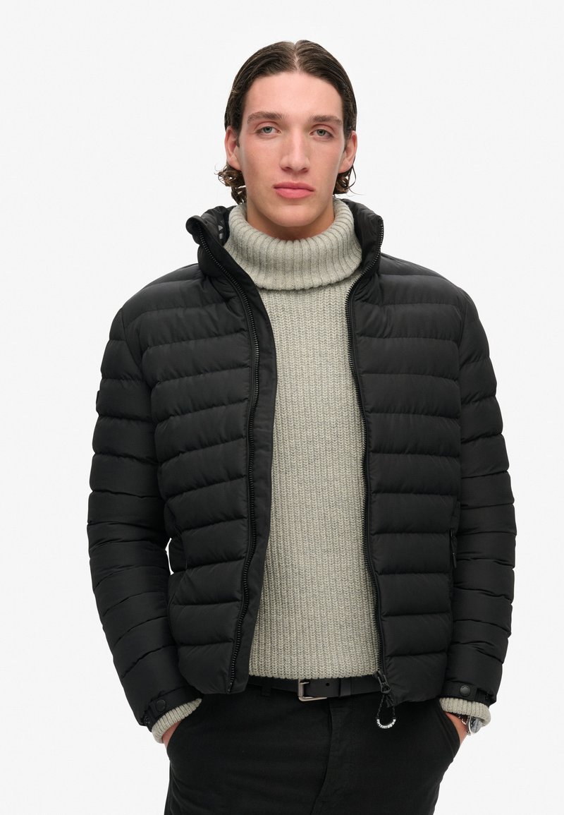 Sort pufferjakke med quiltet mønster, lynlåslukning og hætte. Båret over en lys grå ribbet turtleneck og sorte bukser.