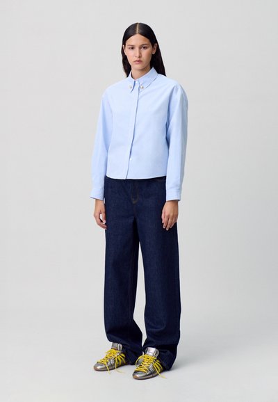 Chemise bleu clair à manches longues, associée à un jean large bleu foncé et des baskets argentées avec des lacets jaunes.