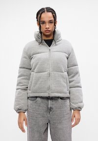 Person med fletninger iført en lys grå pufferjakke og slidt grå jeans, der står foran en ensfarvet hvid baggrund.