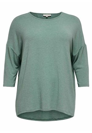 Top vert féminin ample avec manches 3/4 et encolure ronde, ourlet arrière légèrement plus long.