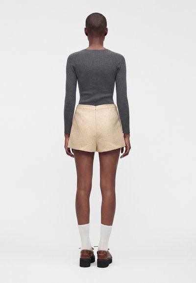 Personne portant un haut gris à manches longues, un short beige taille haute, des chaussettes blanches et des chaussures compensées marron, debout de dos sur fond blanc.