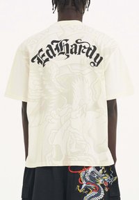 Bílý bavlněný tričko s nápisem "Ed Hardy" černým kurzívou na zádech a jemným designem draka v pozadí. Krátké rukávy.