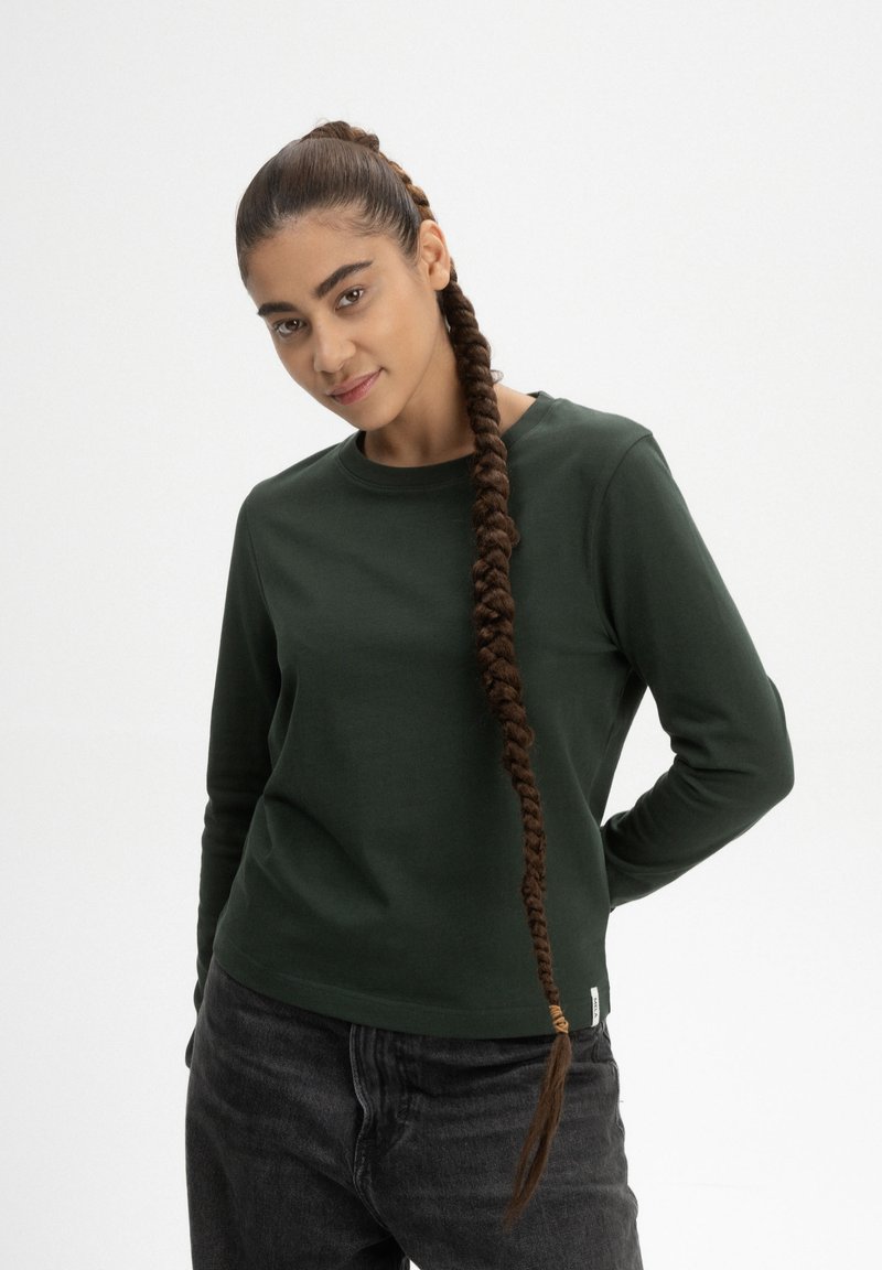 Donkergroene lange mouwen shirt met een ronde hals, gemaakt van zachte stof. Gecombineerd met donkergrijze jeans en een gevlochten kapsel.