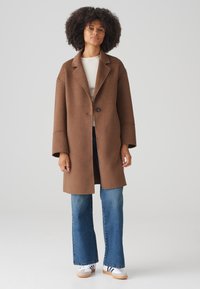 Brown wool blend coat with a notch lapel, single button closure, and wide sleeves, paired with blue flared jeans and white sneakers.