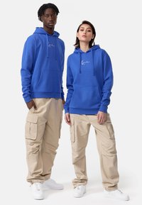 Modre kapuce s sprednjimi žepi in vrvico, v kombinaciji s svetlo bež cargo hlačami z stranskimi žepi in belimi supergami.