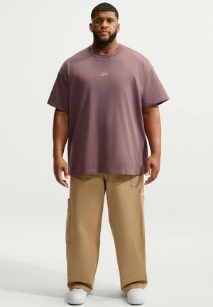 Hombre de pie mirando hacia adelante con una camiseta suelta de Nike color malva, pantalones beige, zapatillas blancas y un collar fino sobre un fondo liso y claro.