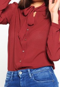 Chemisier bordeaux à boutons avec un col noué, motif texturé, manches longues et détail œillet. Associé à un jean bleu.