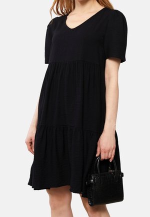 Femme portant une robe noire à manches courtes avec une jupe à volants, tenant un petit sac à main noir texturé dans la main droite.