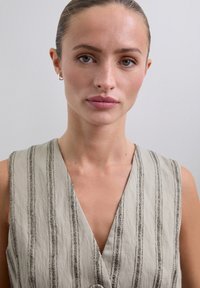 Kvinde med trukket hår iført små hoopsøreringe og en ærmeløs beige top med lodrette striber mod en ensfarvet baggrund.