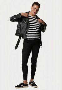 Zwarte leren motorjack over een zwart-wit gestreepte top, gecombineerd met zwarte leggings en zwarte sneakers met beige accenten.