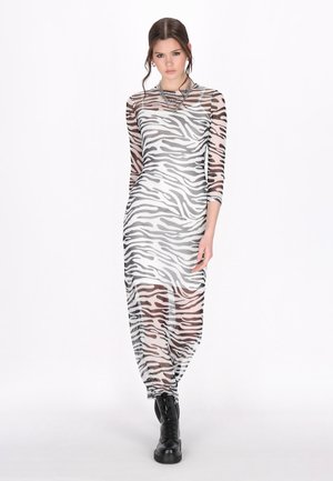 Vrouw die een doorschijnende, lange mouw zebraprint jurk draagt met zwarte laarzen en laagjes zilveren kettingen als accessoires, staand tegen een witte achtergrond.