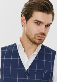 Ciemnoniebieska kamizelka w kratę, wykonana z teksturowanej tkaniny. Posiada jeden guzik oraz małą kieszonkę na piersi. Noszona na białej koszuli.