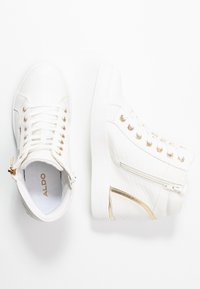 Vita hög-top sneakers med texturerat mönster, guldöglor, sidodragkedjor och guldaccenter, visade från topp- och sidovy på vit bakgrund.