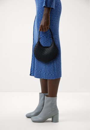 Black woven handbag held by a person, paired with light blue ankle boots and a blue ribbed dress with side slits. Neutral background.
