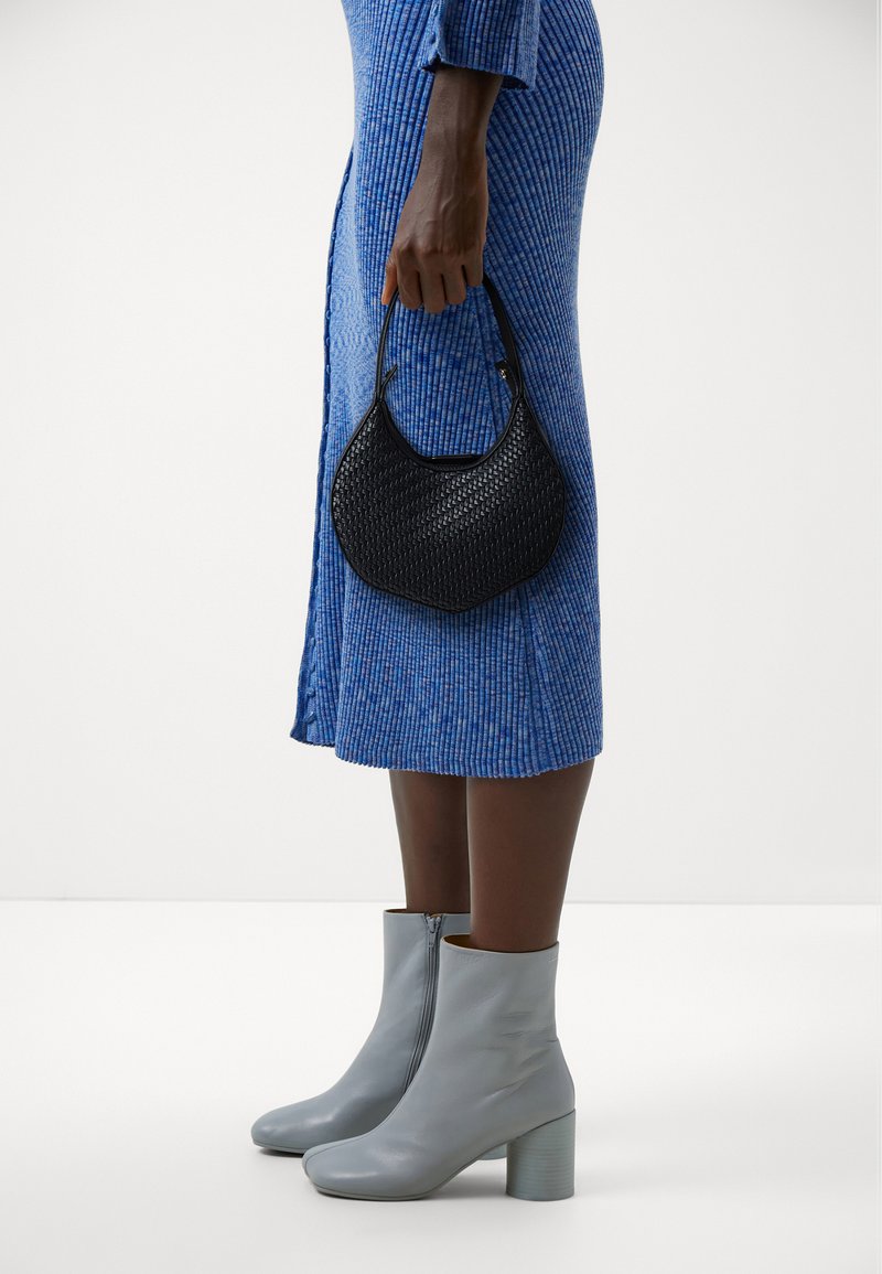 Borsa a tracolla nera intrecciata tenuta da una persona, abbinata a stivaletti azzurri e a un vestito blu a coste con spacco laterale. Sfondo neutro.