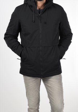 Uomo che indossa una giacca nera con cappuccio, con le mani nelle tasche, e pantaloni grigio chiaro su sfondo bianco.