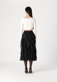 Femme aux longs cheveux noirs portant un crop top blanc à manches longues, une jupe midi en tulle noire superposée, des bas noirs et des chaussures à talons noires, debout de dos.