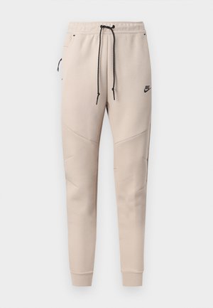 Beige Nike joggebukser med svart snor, glidelåslomme på siden, sydd knepanel og elastiske mansjetter.