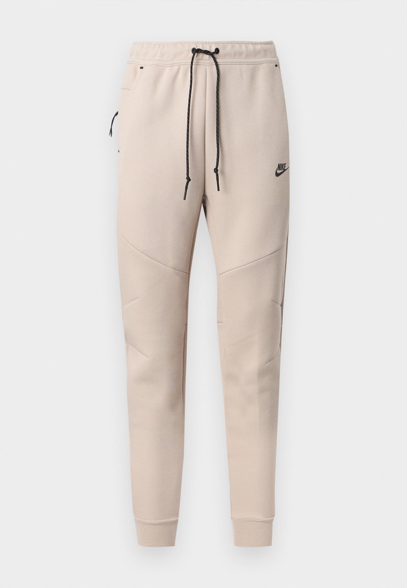Pantalones deportivos beige de Nike con cordón negro, bolsillo lateral con cremallera, paneles de rodilla cosidos y puños elásticos.