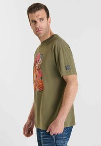 Camiseta de algodón verde oliva con un colorido diseño floral en el frente y un pequeño parche con logo en la manga izquierda.