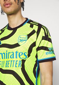 Camiseta de fútbol amarilla neón con rayas negras de cebra, con texto azul y un logo. Hecha de tela transpirable con un cuello redondo.