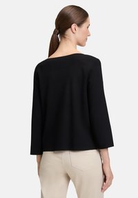 Czarny sweter robiony na drutach z szerokim dekoltem, rękawami o długości trzy czwarte oraz luźnym krojem, w połączeniu z jasnobeżowymi spodniami.