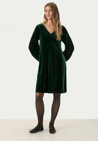 Grünes Samtkleid mit langen Ärmeln, V-Ausschnitt, knielangem Schnitt, glatter Textur, kombiniert mit schwarzen flachen Schuhen und transparenten Strumpfhosen.