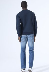 Marineblauwe sweatshirt met een gestructureerd ruitpatroon, gecombineerd met lichtblauwe straight-leg jeans en witte sneakers, gezien vanaf de achterkant.