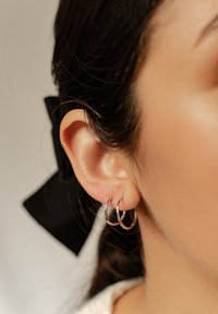 Srebrne kolczyki w kształcie kół noszone są w uchu, charakteryzują się podwójnym wzorem kółek i polerowanym wykończeniem. Włosy są związane czarną wstążką.