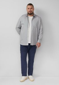Camicia a maniche lunghe con bottoni di colore grigio chiaro con motivo a scacchi, indossata sopra una maglietta bianca, abbinata a pantaloni blu scuro e sneakers bianche.