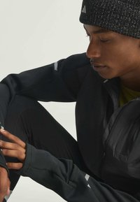 Young person wearing black knit beanie and dark athletic jacket, sitting with arm resting on bent knee against light background.