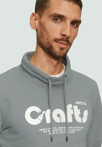 Felpa grigia con cappuccio a collo alto, caratterizzata da una grande stampa bianca con la scritta "Crafts" e dettagli sui materiali artistici. Inclusi i laccetti regolabili.