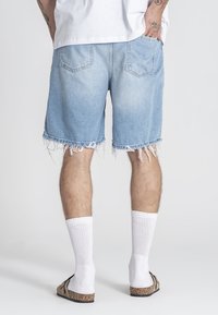 Traper šortsevi svijetloplave boje s razdrapanim rubovima, kombinirani s bijelim čarapama i smeškim sandalama. Vidljive stražnje d žepove i ležeran kroj.