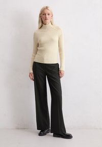 Crèmekleurige geribbelde coltrui, slim fit, gecombineerd met hoge zwarte wijde broek. Model staat tegen een lichte, gestructureerde achtergrond.